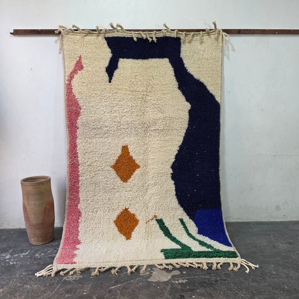 Abstract Azilal Moroccan rug hanging on display, cream wool with navy, pink, green and orange shapes