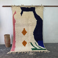Abstract Azilal Moroccan rug hanging on display, cream wool with navy, pink, green and orange shapes