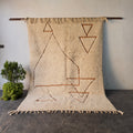 Traditional hand-woven Moroccan Berber rug hanging on display, cream wool with rust triangle and diamond motifs