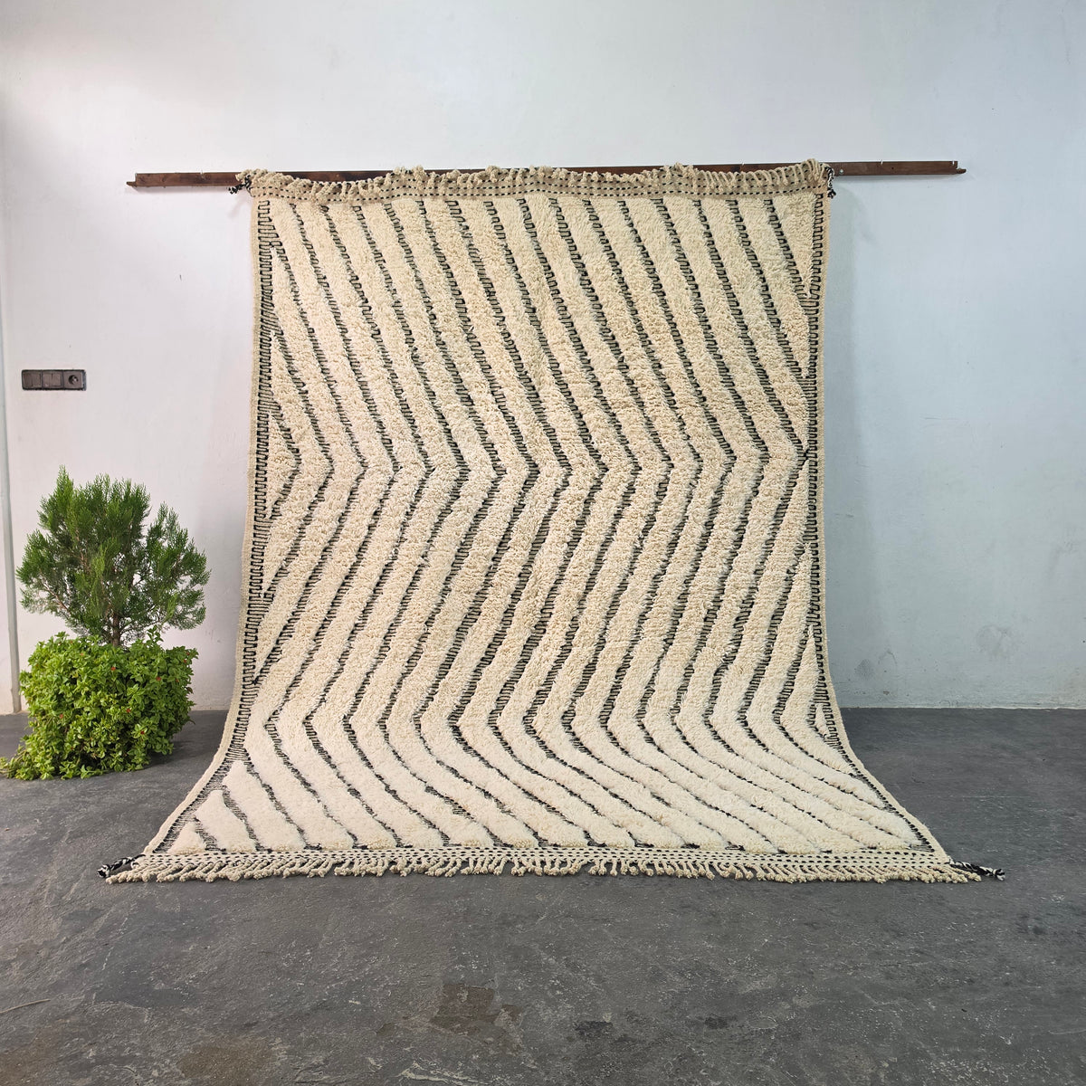 Handmade Moroccan Beni Ourain wool rug hanging on display, ivory wool with black wavy chevron pattern